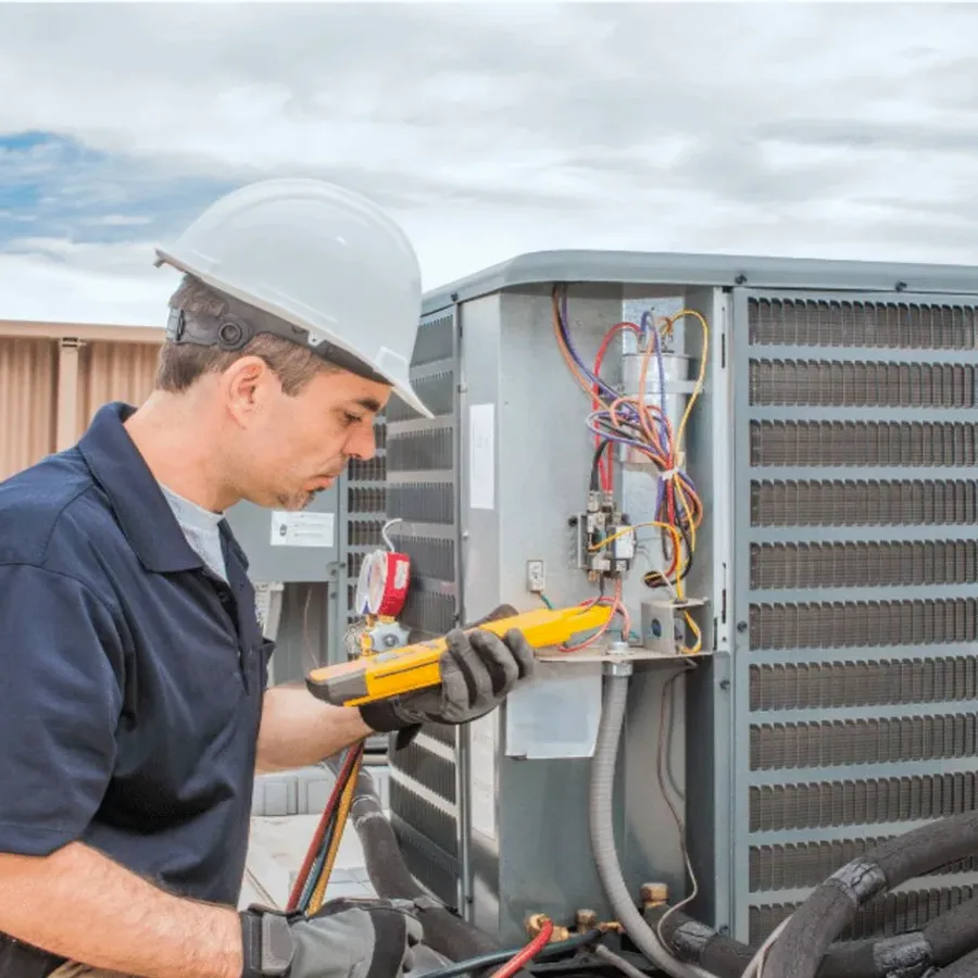 HVAC technician in a hard hat testing an outdoor air conditioner with a multimeter